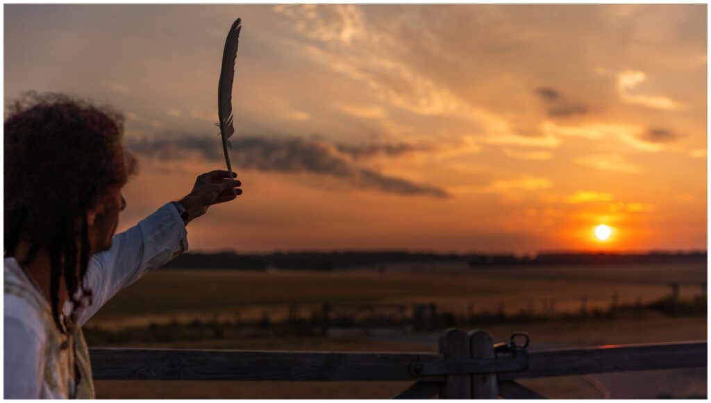 Shaman holding a feather and watching the red sunrise landscape at Stonehenge during a sacred ceremony